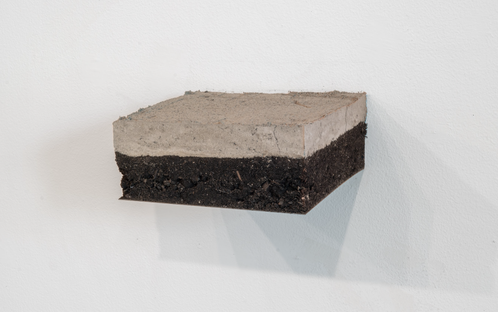 Image: Mariana Noreña, Tierra Ceniza, 2024. Ashes and soil. A gritty, rough grey rectangle, atop a brown rectangle, with clear cut outer layer. It is floating on a white wall. Photo by Jonas Müller-Ahlheim.